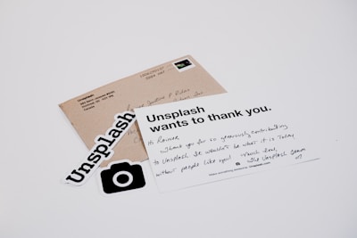 A white card with a handwritten message and printed text reads 'Unsplash wants to thank you.' The card is placed on a table with a brown envelope and Unsplash stickers. The message on the card expresses gratitude, and the envelope has an address label.