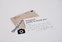 A white card with a handwritten message and printed text reads 'Unsplash wants to thank you.' The card is placed on a table with a brown envelope and Unsplash stickers. The message on the card expresses gratitude, and the envelope has an address label.
