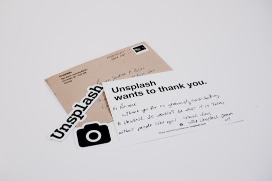 A white card with a handwritten message and printed text reads 'Unsplash wants to thank you.' The card is placed on a table with a brown envelope and Unsplash stickers. The message on the card expresses gratitude, and the envelope has an address label.