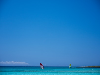 Two windsurfers are gliding over a clear, turquoise sea under a vast blue sky. The horizon is visible in the distance, and there is a small patch of land on the right side of the image. The scene conveys a sense of freedom and leisure.