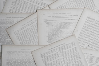 Several sheets of paper with printed text are scattered and overlapping. The text appears to be related to educational topics, with headings like 'Natural and Common Objects' and 'Free-Arm and Industrial Drawing.' The pages are arranged casually, creating a layered effect.