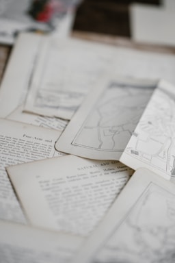 A collection of old papers and documents with printed text and maps laid out overlapping on a surface. Some of the pages have visible text, while others appear to display diagrams or maps.