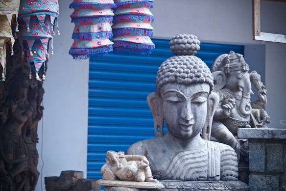 A detailed arrangement of religious sculptures, prominently featuring a serene stone statue of a meditative figure alongside another intricately carved statue. Colorful fabric lamps with geometric designs hang above, adding a vibrant contrast to the monochrome stone figures. A textured, blue metal shutter serves as the backdrop, enhancing the visual depth.
