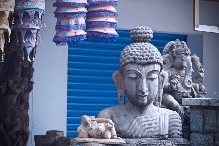 A detailed arrangement of religious sculptures, prominently featuring a serene stone statue of a meditative figure alongside another intricately carved statue. Colorful fabric lamps with geometric designs hang above, adding a vibrant contrast to the monochrome stone figures. A textured, blue metal shutter serves as the backdrop, enhancing the visual depth.