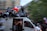 A large group of people are gathered on and around a moving van. Many of them are waving or holding a flag with blue, white, and red stripes. The scene appears to be festive and energetic, with some people sitting on top of the van and others standing or cheering. The background is blurred, indicating motion.