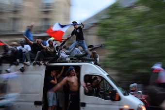 A large group of people are gathered on and around a moving van. Many of them are waving or holding a flag with blue, white, and red stripes. The scene appears to be festive and energetic, with some people sitting on top of the van and others standing or cheering. The background is blurred, indicating motion.