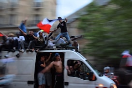 A large group of people are gathered on and around a moving van. Many of them are waving or holding a flag with blue, white, and red stripes. The scene appears to be festive and energetic, with some people sitting on top of the van and others standing or cheering. The background is blurred, indicating motion.