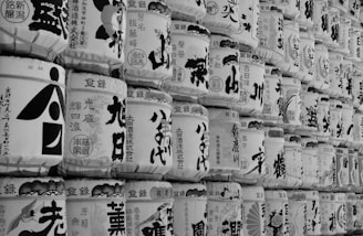 A close-up view of stacked traditional Japanese sake barrels, each adorned with various intricate calligraphic characters and illustrations. The barrels are arranged in multiple rows, creating a uniform pattern that highlights the craftsmanship and cultural significance of the items.
