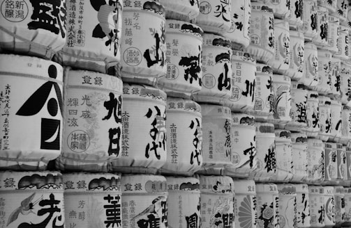 A close-up view of stacked traditional Japanese sake barrels, each adorned with various intricate calligraphic characters and illustrations. The barrels are arranged in multiple rows, creating a uniform pattern that highlights the craftsmanship and cultural significance of the items.