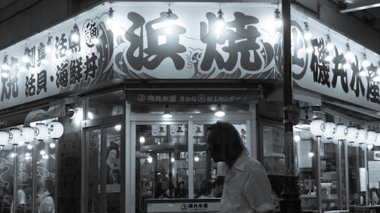A black-and-white photograph of a Japanese restaurant, with prominent signage featuring Japanese text. Lanterns and lights illuminate the entrance. A person in the foreground has their back turned towards the camera, adding a sense of anonymity and interaction with the environment.