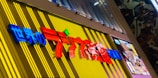 A colorful store sign with Japanese characters, prominently displaying vibrant red and blue text against a yellow and striped background. On the right side, there is a digital screen displaying content with anime-style artwork. The surrounding area has a modern and urban setting with architectural elements including a metal canopy and window panes.