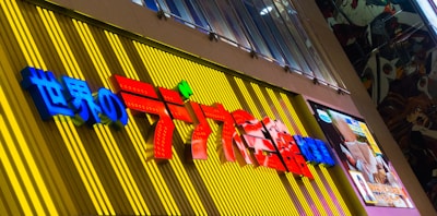 A colorful store sign with Japanese characters, prominently displaying vibrant red and blue text against a yellow and striped background. On the right side, there is a digital screen displaying content with anime-style artwork. The surrounding area has a modern and urban setting with architectural elements including a metal canopy and window panes.