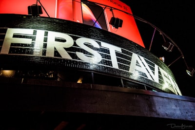 A prominent black building facade with large white letters spelling 'FIRST AVENUE' is illuminated by external lights. Above it, a vibrant red billboard, partially visible, adds contrast to the scene.