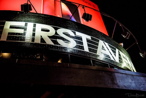 A prominent black building facade with large white letters spelling 'FIRST AVENUE' is illuminated by external lights. Above it, a vibrant red billboard, partially visible, adds contrast to the scene.