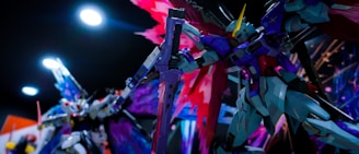 Colorful and intricately designed robotic figures are posed in a dynamic stance, with one in the foreground wielding a large gun and another in the background seemingly engaged in action. The backdrop is filled with vibrant and abstract light patterns, adding energy to the scene.