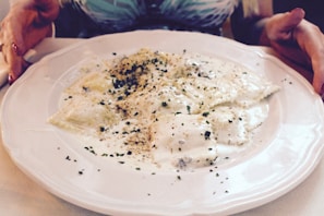 A plate of ravioli covered in a creamy white sauce, sprinkled with herbs and spices, is placed on a table. Two hands frame the dish, suggesting it is ready to be served or enjoyed.