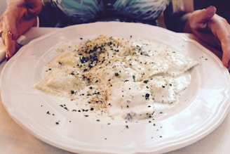 A plate of ravioli covered in a creamy white sauce, sprinkled with herbs and spices, is placed on a table. Two hands frame the dish, suggesting it is ready to be served or enjoyed.