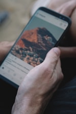 A hand is holding a smartphone displaying a social media app with a photo of a mountainous landscape.