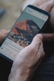 A hand is holding a smartphone displaying a social media app with a photo of a mountainous landscape.