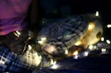 A person wearing plaid pajamas holds a string of glowing fairy lights in a dimly lit room, creating a cozy atmosphere. A stuffed animal is partially visible next to them.