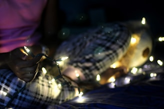 A person wearing plaid pajamas holds a string of glowing fairy lights in a dimly lit room, creating a cozy atmosphere. A stuffed animal is partially visible next to them.