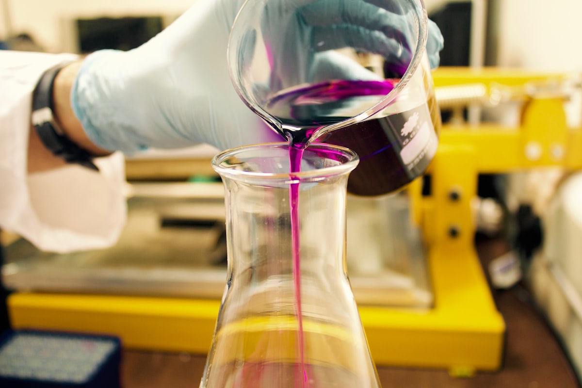 a gloved hand wearing a watch in a lab pouring a purple liquid from a glass beaker into another glass beaker.