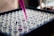 A close-up view of a tray filled with transparent laboratory test tubes, some of which are being filled with a pink liquid using a pipette. The setting appears to be a laboratory environment focused on scientific experiments or medical testing.