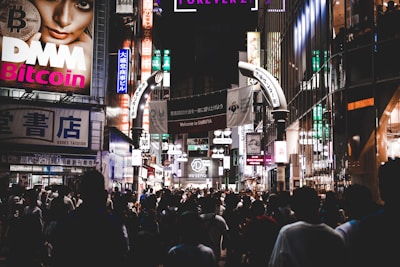 A bustling city scene at night filled with a crowd of people walking through brightly lit streets adorned with numerous commercial billboards and neon signs. The environment is vibrant with various advertisements and shop signs in different languages, contributing to an urban and lively atmosphere.