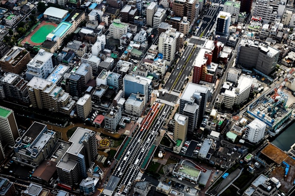Aerial view of a densely packed urban area featuring a mix of high-rise buildings, residential structures, and commercial facilities. The streets are bustling with cars, and there is a noticeable presence of green sports fields and construction sites. The layout suggests a well-organized cityscape with diverse architecture.