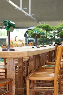 Several wooden chairs are arranged around a long wooden table with green glass lamps evenly spaced along the surface. The setting appears to be indoors with natural light streaming in, complemented by lush greenery visible in the background. The ambiance suggests a cozy, communal space.