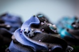 A close-up view of a cupcake with swirled frosting in shades of blue and black. The frosting is intricately decorated with small sugar stars and tiny round sprinkles. The focus is on the central cupcake, with blurred cupcakes in the background.