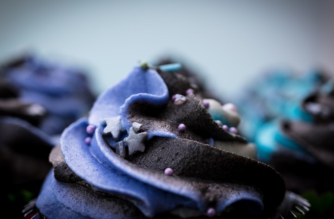 A close-up view of a cupcake with swirled frosting in shades of blue and black. The frosting is intricately decorated with small sugar stars and tiny round sprinkles. The focus is on the central cupcake, with blurred cupcakes in the background.