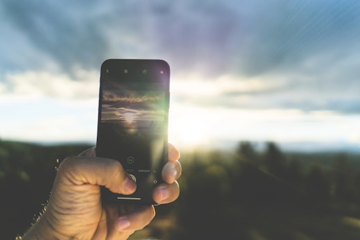 A hand holding a smartphone with the camera app open, capturing a scenic view of a sunset or sunrise. The background features a bright sun partially obscured by clouds, casting soft light across a landscape.