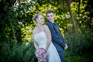 A couple in wedding attire stands back-to-back in a lush, green outdoor setting. The bride is wearing a strapless white gown and holding a bouquet of pink flowers. The groom is dressed in a formal dark suit, smiling at the bride. The background is filled with greenery and trees, creating a serene and romantic atmosphere.
