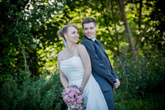 A couple in wedding attire stands back-to-back in a lush, green outdoor setting. The bride is wearing a strapless white gown and holding a bouquet of pink flowers. The groom is dressed in a formal dark suit, smiling at the bride. The background is filled with greenery and trees, creating a serene and romantic atmosphere.