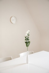 A minimalistic interior with a white table and two chairs. A simple white vase with green foliage and a few white flowers stands in the center of the table. A round wall clock is mounted on the light-colored wall, adding to the clean and serene atmosphere.