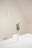 A minimalistic interior with a white table and two chairs. A simple white vase with green foliage and a few white flowers stands in the center of the table. A round wall clock is mounted on the light-colored wall, adding to the clean and serene atmosphere.