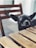 A small black dog, likely a Chihuahua, is peering over a wooden table with its tongue slightly out. The setting is outdoors with a wooden chair in the background.