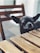 A small black dog, likely a Chihuahua, is peering over a wooden table with its tongue slightly out. The setting is outdoors with a wooden chair in the background.