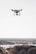 A drone hovering above a blurred distant landscape, featuring a camera mounted underneath its body against a clear sky.