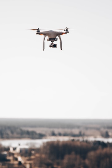A drone hovering above a blurred distant landscape, featuring a camera mounted underneath its body against a clear sky.