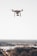 A drone hovering above a blurred distant landscape, featuring a camera mounted underneath its body against a clear sky.