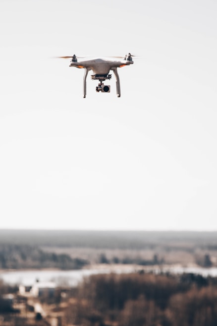 A drone hovering above a blurred distant landscape, featuring a camera mounted underneath its body against a clear sky.