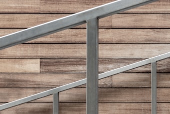 A staircase railing composed of silver metal bars is positioned diagonally against a backdrop of wood paneling. The wood panels have a rustic, natural appearance with various shades of brown, adding a warm contrast to the cool tone of the metal.