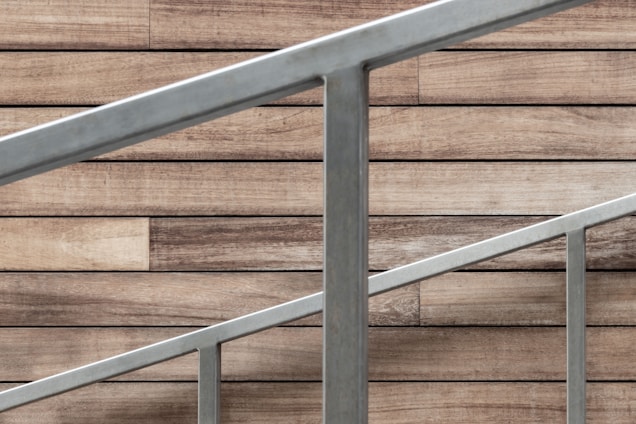 A staircase railing composed of silver metal bars is positioned diagonally against a backdrop of wood paneling. The wood panels have a rustic, natural appearance with various shades of brown, adding a warm contrast to the cool tone of the metal.