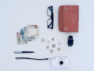 A neatly arranged collection of everyday items on a light-colored surface. These items include a stack of banknotes and several coins, a pair of black glasses, a brown leather wallet, a smartwatch with a black strap, a metallic pen, and a white Nikon camera.