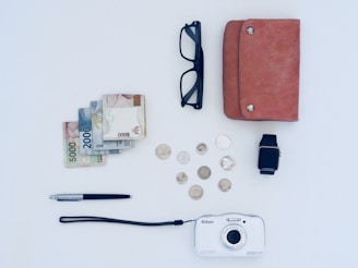 A neatly arranged collection of everyday items on a light-colored surface. These items include a stack of banknotes and several coins, a pair of black glasses, a brown leather wallet, a smartwatch with a black strap, a metallic pen, and a white Nikon camera.