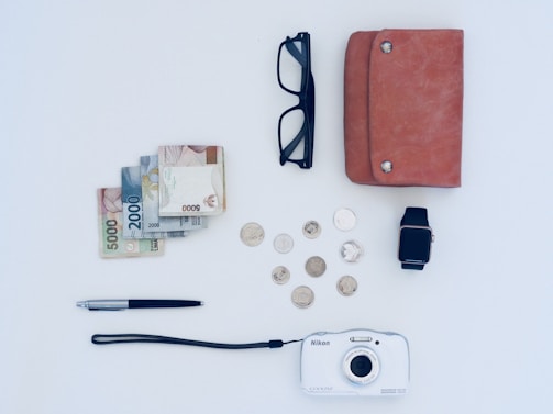 A neatly arranged collection of everyday items on a light-colored surface. These items include a stack of banknotes and several coins, a pair of black glasses, a brown leather wallet, a smartwatch with a black strap, a metallic pen, and a white Nikon camera.