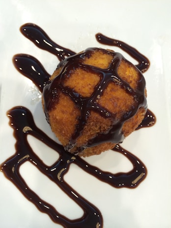 A golden-brown, crispy round dessert is drizzled with rich, dark chocolate sauce, elegantly presented on a white plate.