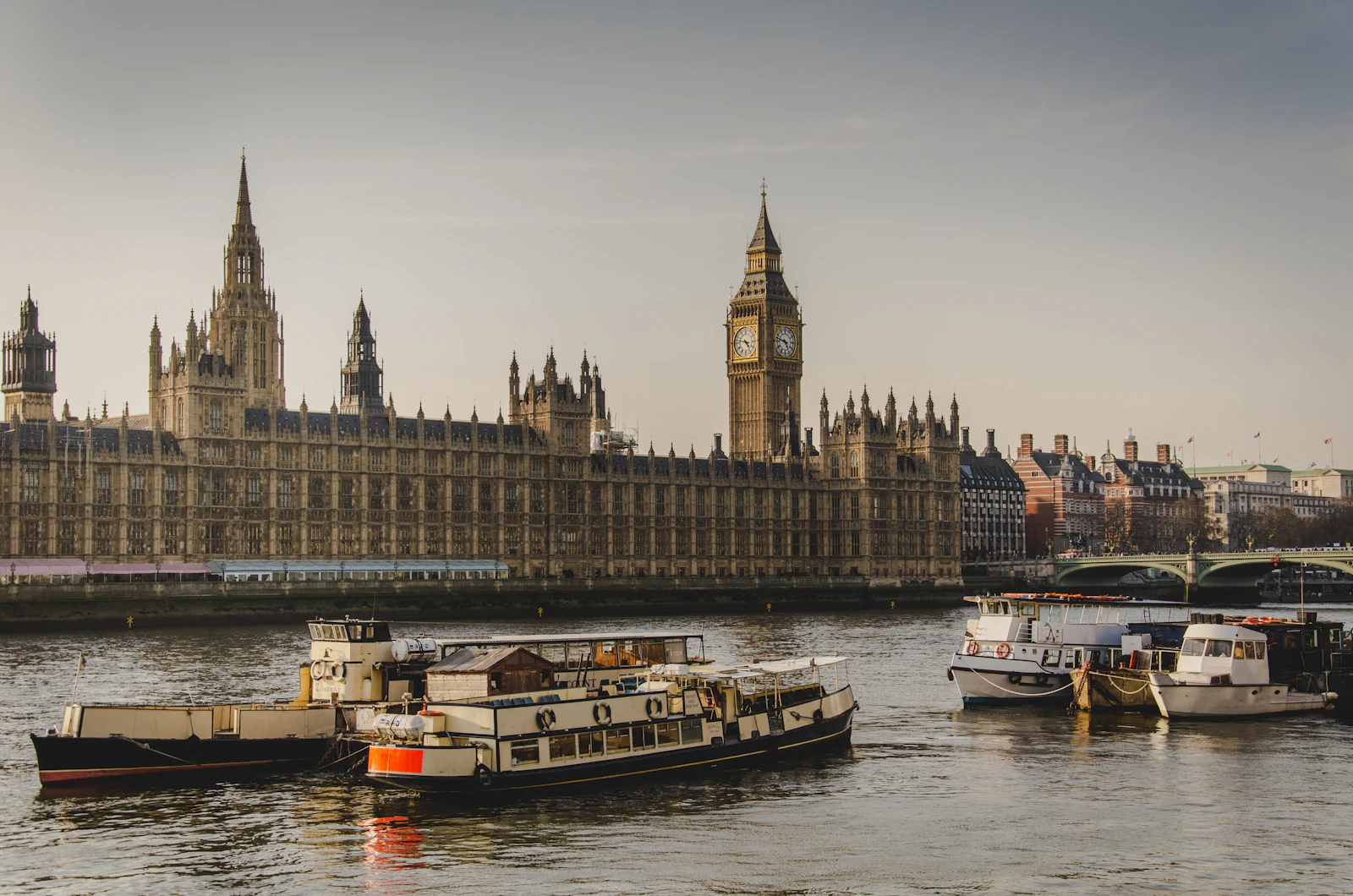 Photograph of Passenger boats move along the Thames in front of the long Gothic river façade of Parliament.